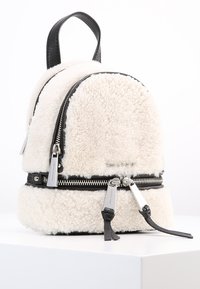 Vit shearling ryggsäck med svarta detaljer, med dubbla dragkedjor och ett handtag på toppen. Rundad form, mjuk textur och framträdande sömmar synliga.