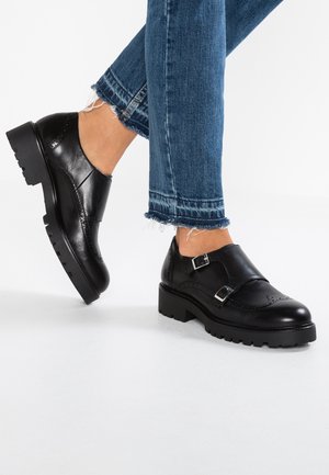 Mustat nahkaiset kaksinkertaiset monkki-solki kengät, joissa on brogue-yksityiskohdat, käytetty kuluneen lahkeen sinisten farkkujen kanssa, kuvattuna sivulta valkoisella taustalla.