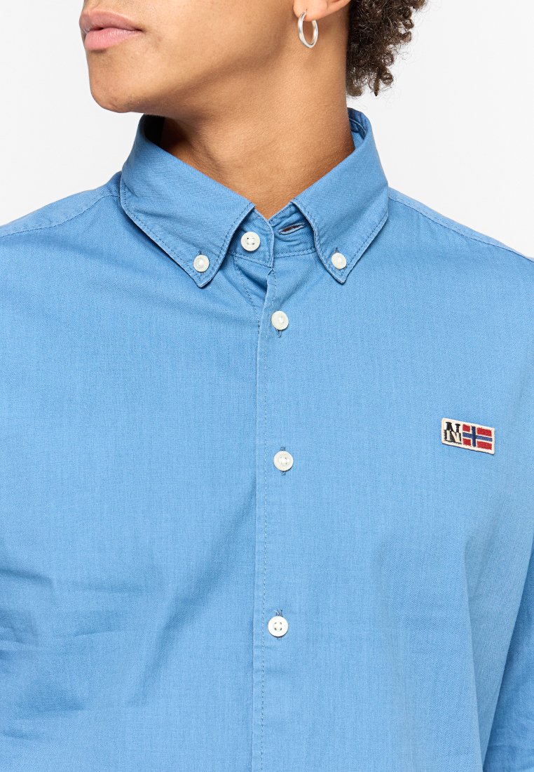 Helles blaues Hemd mit Kragen, mit weißen Knöpfen und einem kleinen gestickten Patch in Rot und Weiß.