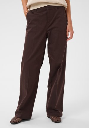 Personne portant un pantalon large marron foncé et des chaussures tissées marron, avec une main dans la poche et un haut neutre visible à la taille.