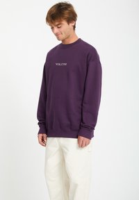 Sweatshirt violet en coton avec un col rond et des poignets côtelés. Présente un logo "VOLCOM" grisé brodé sur la poitrine. Associé à un pantalon blanc.