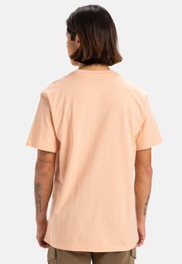 Camiseta de algodón melocotón claro con mangas cortas, cuello redondo y un corte relajado. La vista trasera muestra una textura suave y sin marcas visibles.