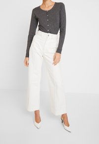 Kvinna i en mörkgrå knappad cardigan med vita högmidjade vida byxor och vita spetsiga klackar, som står framför en neutral bakgrund.