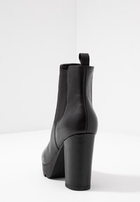 Botte chelsea en cuir noir avec un talon épais et haut, panneaux latéraux élastiques et languette à l'arrière, sur une surface et un fond blanc.