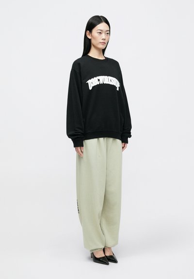 Melnā treniņjakā ar baltu tekstu, gaiši zaļās biksēs ar elastīgām aprocēm un melnās šaurās kurpēs, kas tiek nēsātas stāvoša modeļa.