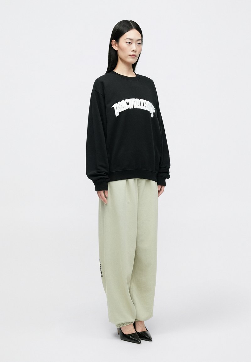 Zwarte sweatshirt met witte tekst, lichtgroene trainingsbroek met elastische boorden, en zwarte pumps, gedragen door een staande model.