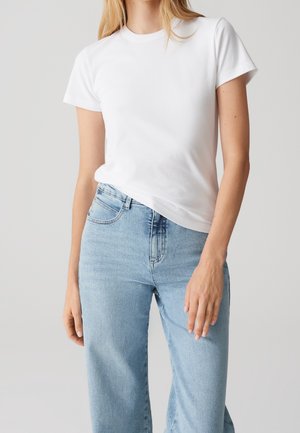 Woman wearing a plain white short-sleeve t-shirt tucked into light blue high-waisted jeans against a neutral background.