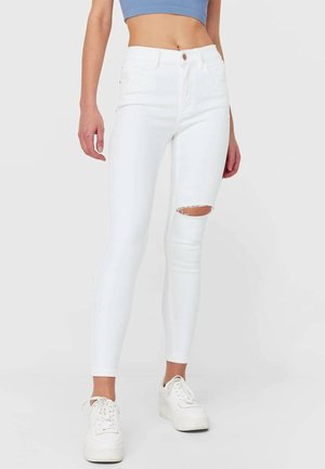 Frau trägt weiße hochtaillierte Skinny-Jeans mit Knieriss, weiße Plateau-Sneaker und ein blaues bauchfreies Oberteil vor einem einfarbigen Hintergrund.