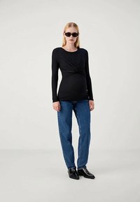 Έγκυος γυναίκα που φοράει μαύρη μπλούζα με μακριά μανίκια και δεσίματα, μπλε τζιν, μαύρα παπούτσια και γυαλιά ηλίου, όρθια μπροστά σε απλό ανοιχτόχρωμο φόντο.