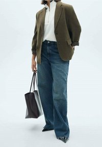 Personne portant un blazer olive surdimensionné, un pull zippé blanc, un jean à jambes larges, des chaussures noires pointues, tenant un grand sac fourre-tout noir.