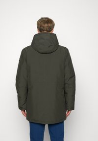 Veste à capuche vert foncé avec une texture lisse. Présente une coupe droite et un ourlet arrière plus long. Design minimaliste, sans motifs visibles.