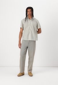 Kortermet mønstret skjorte med lys kremfarge og grønne geometriske former, parret med lyse denimjeans og brune sko.
