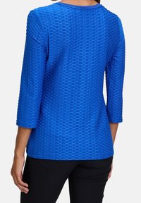 Niebieska, teksturowana bluzka z rękawem o długości trzech kwart, okrągłym dekoltem i wypukłym geometrycznym wzorem na całej powierzchni materiału.