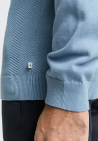 Hellblaues Sweatshirt mit strukturiertem Fischgrätenmuster, gerippten Bündchen und einem kleinen Logosticker an der Seite. Weiches Strickmaterial.