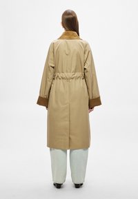 Beež värvi trentšmantel pruuni veluurkraega, nööbitud vöökohaga ja laiemate varrukatega, kombineeritud helesiniste lahtiste teksadega.