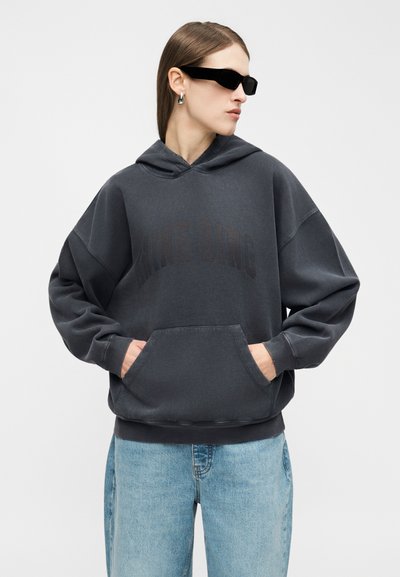 Femme portant un sweat à capuche noir surdimensionné avec poche kangourou, jean bleu clair, lunettes de soleil noires étroites et boucles d'oreilles argentées en créole, regardant vers la droite.