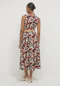 Woman wearing sleeveless, floral-print midi dress with a beige belt facing away, paired with beige heeled mules, standing against white background.