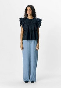 Schwarze Bluse mit gerüschten, gewellten Ärmeln, gestuftem Oberteil und rundem Ausschnitt. Kombiniert mit hellblauen, weit geschnittenen Jeans und schwarzen, spitzen Schuhen.