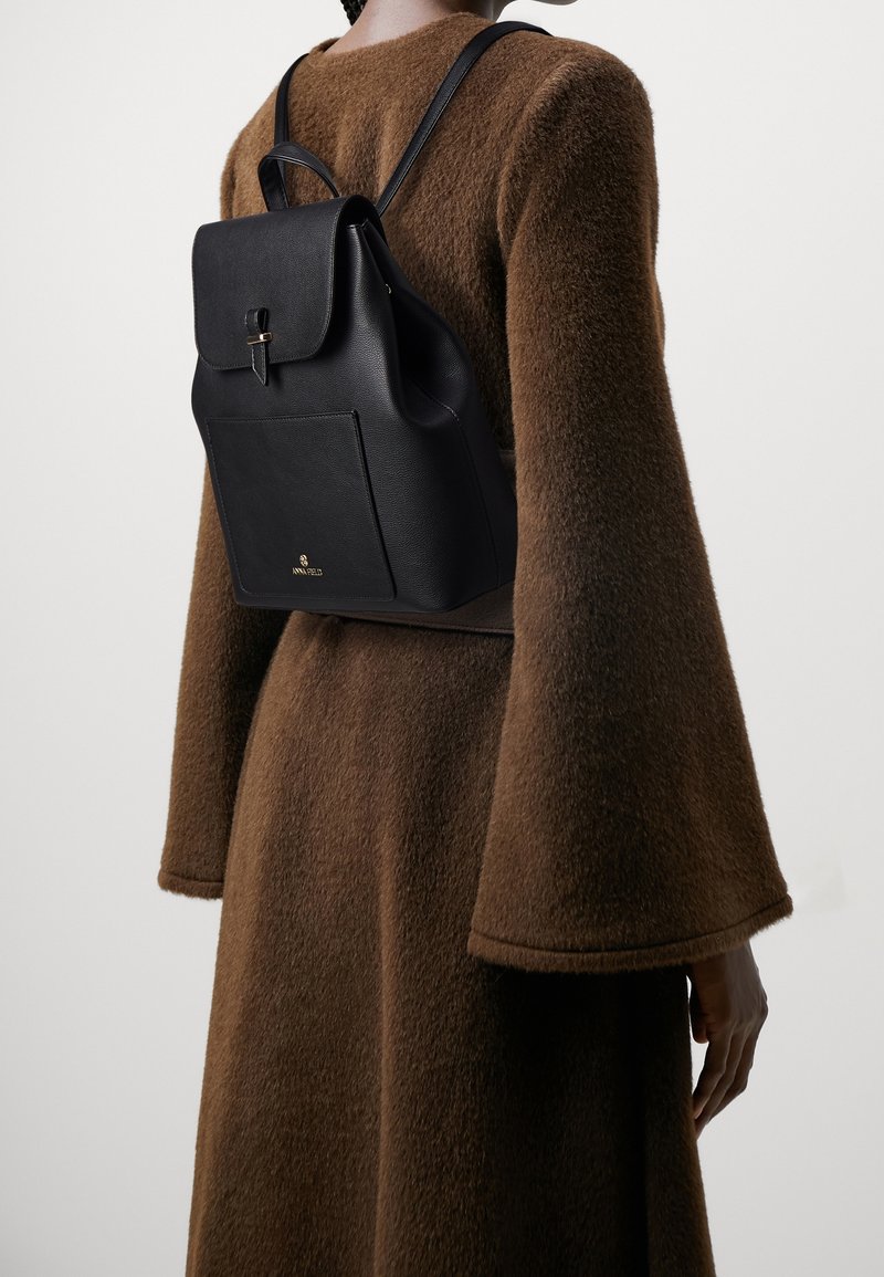 Personne portant un manteau marron texturé à manches larges et un petit sac à dos en cuir noir avec une poche avant et une fermeture à rabat.