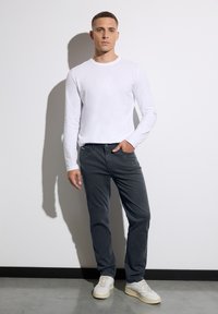 Valkoinen pitkähihaisen paidan, harmaat housut ja valkoiset lenkkarit. Sileä kangas, slim fit. Malli seisoo valkoista seinää vasten, varjo heitetty.