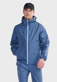 Giacca impermeabile blu con cappuccio, chiusura frontale con zip, polsini elastici e tasche laterali con zip. Abbinata a pantaloni azzurri chiari.