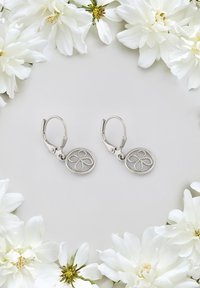 Silverörhängen med ett blommigt utskärningsdesign. Örhängena hänger från en låsback med en polerad yta, mot en bakgrund av vita blommor.