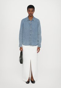 Denim button-up shirt in light blue, paired with a white maxi skirt featuring a front slit, and a black textured handbag. Black flats complete the look.