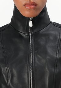 Giubbotto in pelle nera con colletto alto e dettagli in zip argentata. Presenta una texture liscia e una vestibilità su misura, con accenti di cuciture visibili.