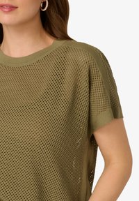 Zeleno-olivový pletený top s krátkým rukávem a volným, otevřeným pletením. Má žebrovaný výstřih a hladké boční akcenty.