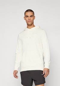 Felpa crema con tasca a marsupio, cappuccio con cordino e polsini a costine. Presenta un piccolo logo bianco sul petto. Indossata con pantaloni corti scuri.