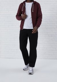 Muž v burgundské mikině na zip přes bílém tričku, černých kalhotách a stříbrných teniskách stojící proti bílé cihlové zdi.