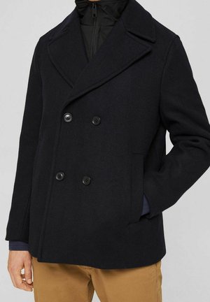 Czarny dwurzędowy płaszcz typu pea coat z szerokimi klapami, noszony na czarnej kurtce na zamek błyskawiczny i beżowych spodniach, ręka w kieszeni, na jednolitym tle.