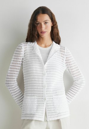 Ung kvinde med langt bølget hår iført en hvid tekstureret knapcardigan over en hvid top, hænderne bag ryggen, neutralt baggrund.