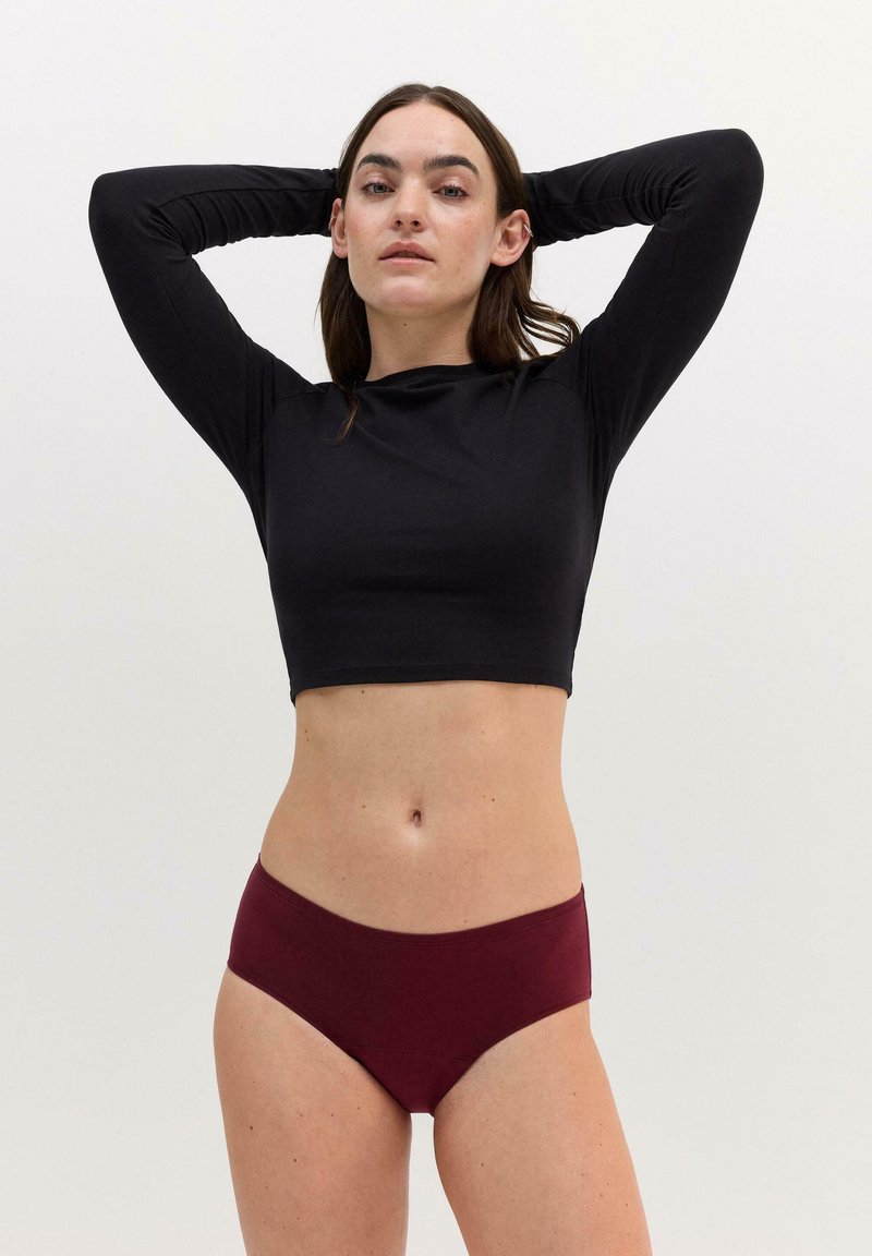 Frau in einem schwarzen, langärmeligen Cropped-Top und burgunderfarbenem Unterwäsche, die mit den Händen hinter dem Kopf vor einem schlichten weißen Hintergrund steht.