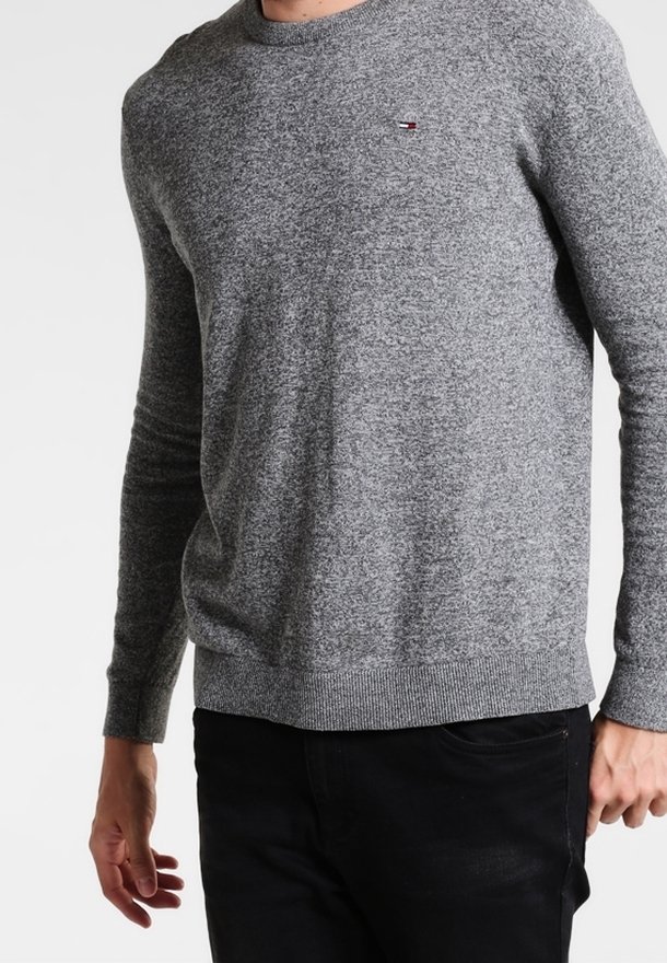 Homme portant un pull gris ajusté à col rond avec un petit logo sur la poitrine et un pantalon noir, debout devant un fond clair uni.