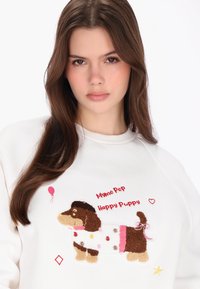 Fehér pulóver, barna és bézs színű hímzett kutyával, rózsaszín léggömbbel és pöttyökkel díszítve, piros és csillag motívumokkal kiegészítve.