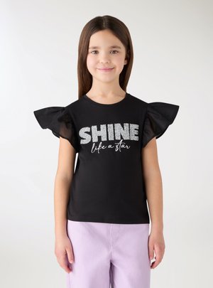 Ragazza con lunghi capelli castani indossa una maglietta nera con maniche a sbuffo e la scritta "SHINE like a star", abbinata a pantaloni viola chiaro.