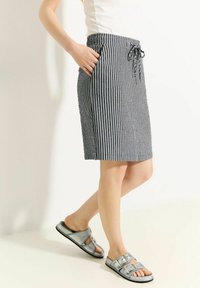 Gestreifte knielange Rock in Schwarz und Weiß, mit einem elastischen Bund und einem Kordelzug sowie seitlichen Taschen. Getragen mit metallischen Sandalen.
