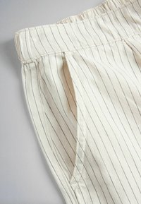 Prim-plan cu pantaloni albi cu dungi verticale subțiri negre și buzunar lateral pe un fundal gri deschis.