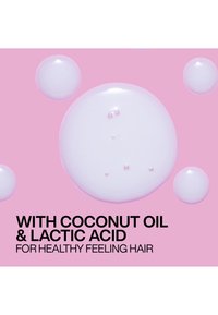 Gouttes de liquide blanc brillant sur fond rose avec un texte indiquant huile de coco et acide lactique pour des cheveux qui ont une sensation de santé.