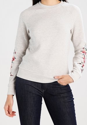 Jasnoszara bluza z różowym kwiatowym haftem na rękawach, noszona z ciemnoniebieskimi dżinsami, ręka oparta w kieszeni.
