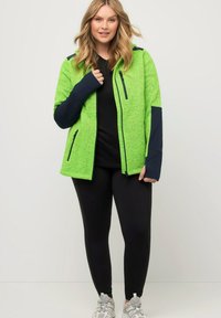 Giacca in pile verde con accenti blu navy, tasche con zip e cappuccio. Indossata sopra una maglietta nera e abbinata a leggings neri e sneakers.