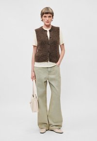 Colete castanho felpudo sobre uma camisa creme, combinado com calças largas verde claro. O modelo segura uma bolsa estruturada creme.