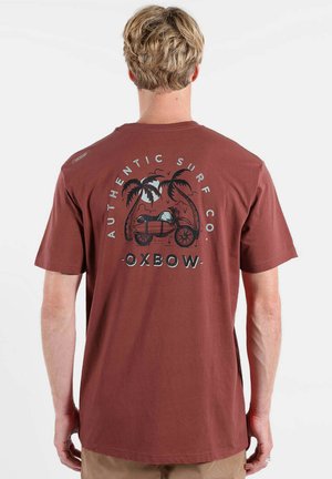 Homme aux cheveux blonds portant un t-shirt marron avec un palmier, une moto et le texte « Authentic Surf Co Oxbow » dans le dos.