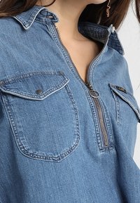 Kvinna i blå denimskjorta med dragkedja fram, fickor med lock och knappar, samt krage, som visar en del av hennes nacke och örhängen.