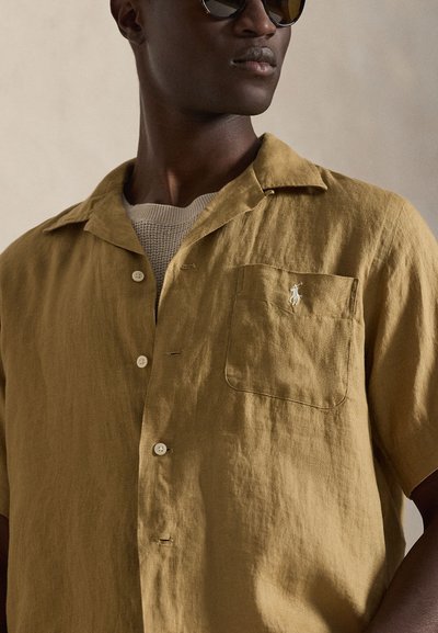 Hombre con camisa de manga corta color beige con bolsillo en el pecho y logo blanco bordado, camiseta beige de punto debajo y gafas de sol oscuras