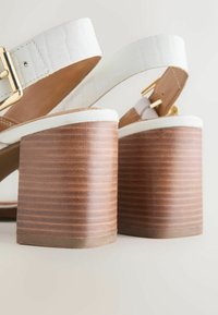 Sandales blanches en cuir à talons blocs, avec brides à boucle dorée et talons en bois empilés sur un fond clair.