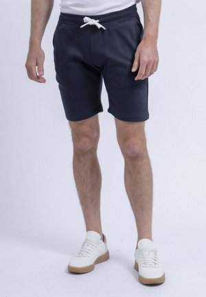 Homme portant un short bleu marine avec cordon et des baskets blanches, debout sur un fond clair uni.