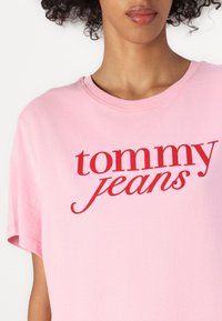 Kvinne som har på seg en lys rosa T-skjorte med rødmf "tommy jeans" tekst trykt over brystet, krøllete hår delvis synlig.