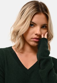 Tmavě zelený svetr s výstřihem do v a dlouhými rukávy, vyrobený z měkkého materiálu. Detailní pohled zdůrazňuje strukturu a detaily výstřihu.